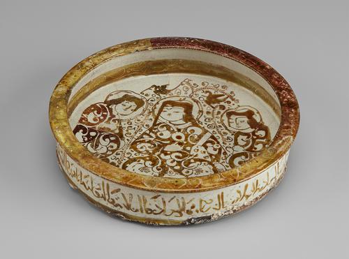 Shallow, decorated dish. The inside is decorated with three female figures in patterned robes, with swirling decoration around them. The exterior of the dish is inscribed with calligraphy, and the rim is decorated in yellow-brown with faint swirling marks.