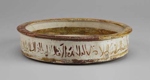 Side view of a shallow, decorated dish. The sides are decorated with a calligraphic inscription in yellow-brown, with the rim coloured solid yellow-brown. Similar solid yellow-brown striping is visible inside the dish and at the bottom of the side.