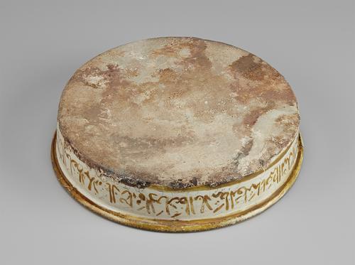 Underside of a shallow, decorated dish. The bottom is unglazed and mottled. The decorated exterior rim is visible, showing the yellow-brown calligraphy upside down, with the edge of the dish coloured solid yellow-brown.