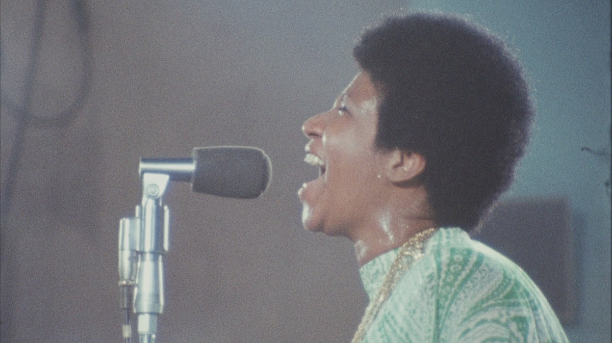 A young Aretha Franklin, sweating and wearing a green dress, sings passionately into a microphone.