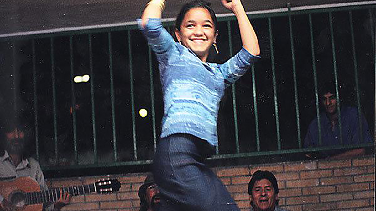A girl wearing a blue shirt throws her hands up in a dance move, as men sing and play guitar in the background.