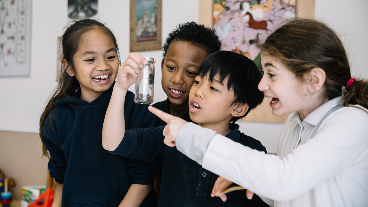 A child holds up a specimen jar while another child points at it and two more children look on smiling.