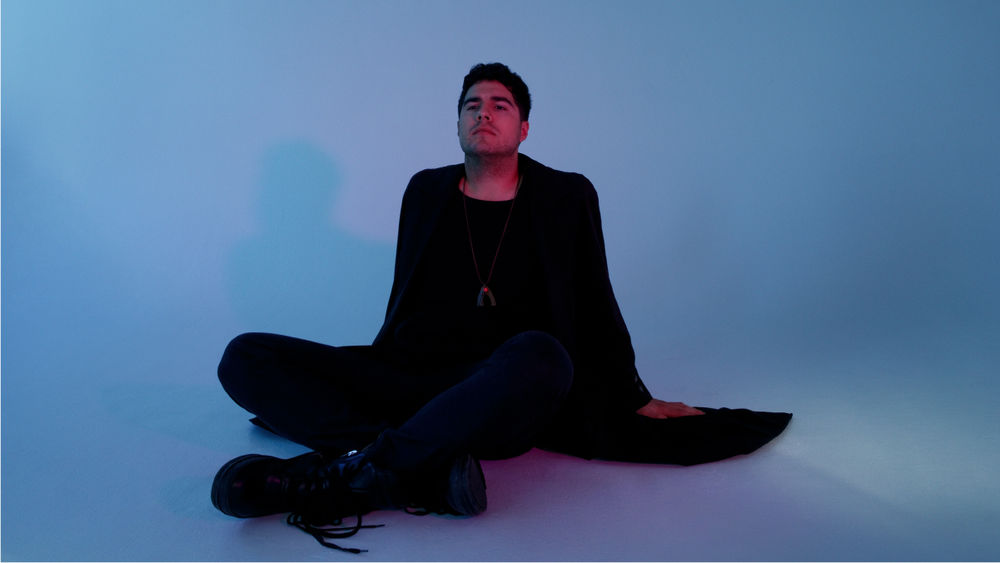 Against a blue backdrop and floor, Jeremy Dutcher sit cross-legged and leans back on his arms, looking upwards.