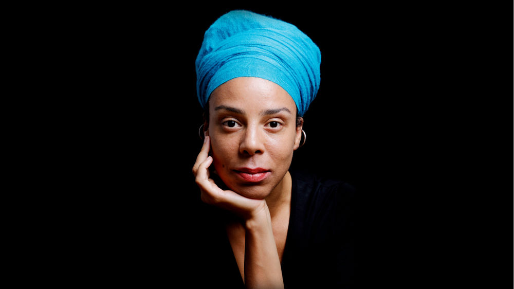 Tanya Evanson, wearing a blue turban, poses with her chin on her hand, against a black backdrop.
