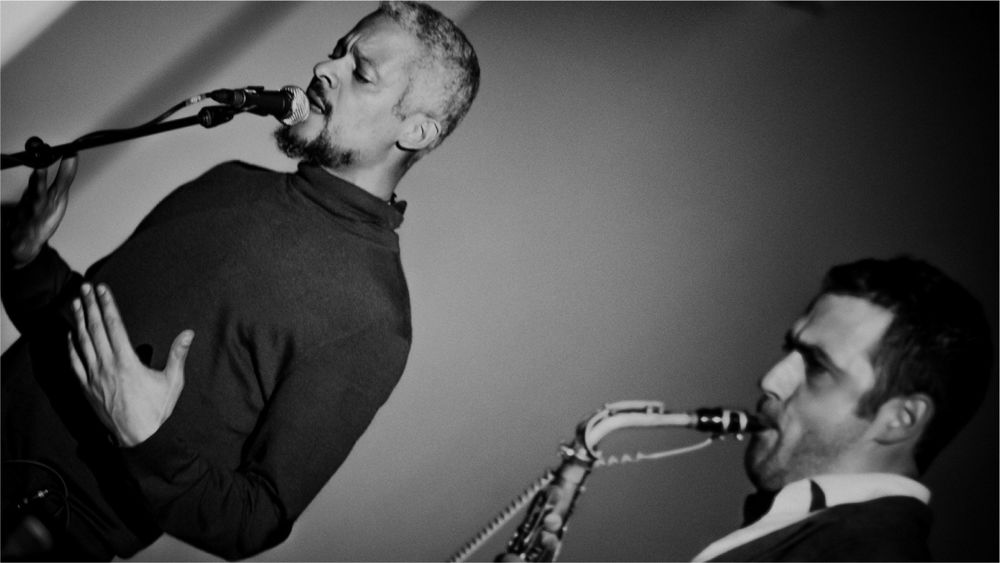 A black and white image of Kaie Kellough singing into a mic and Jason Sharp playing sax.