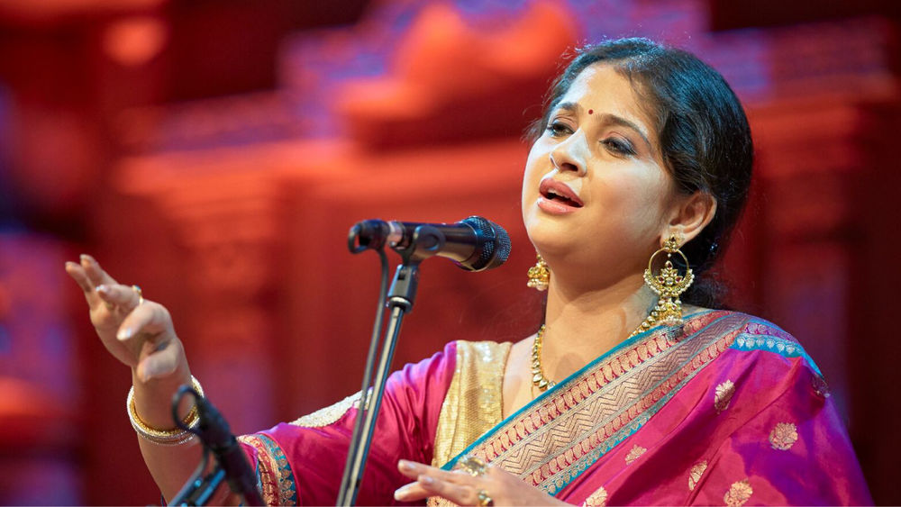 Kaushiki Chakraborty, wearing a magenta and gold shalwar kameez, sings on stage in front of a microphone against a red backdrop.