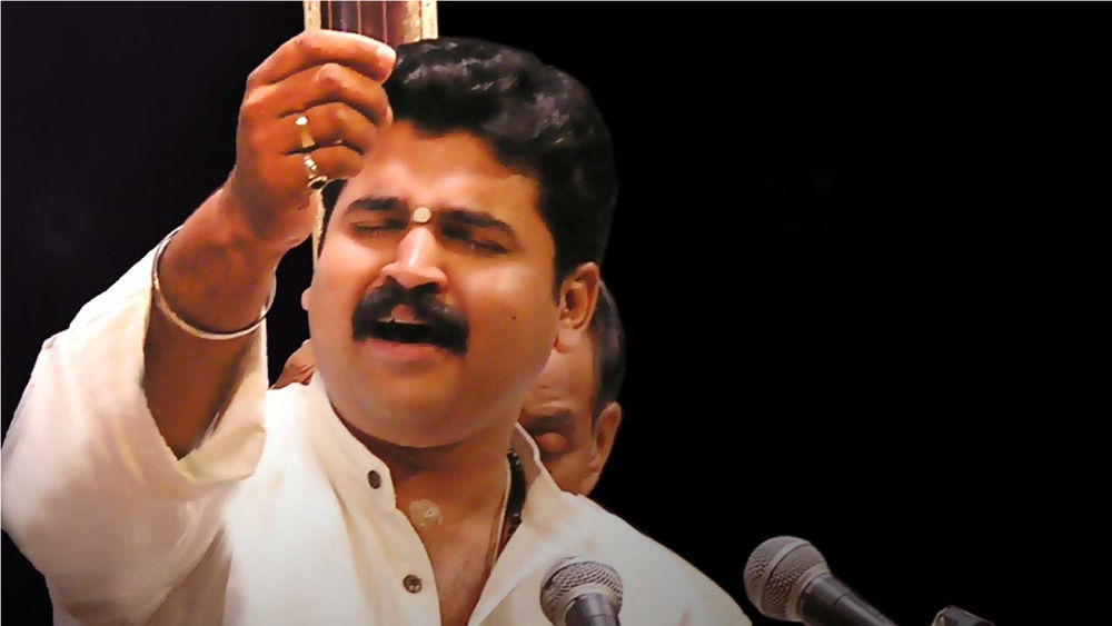 Bhuvanesh Mukul Komkali , wearing a white button-up shirt, sings into a microphone with eyes closed against a black backdrop.