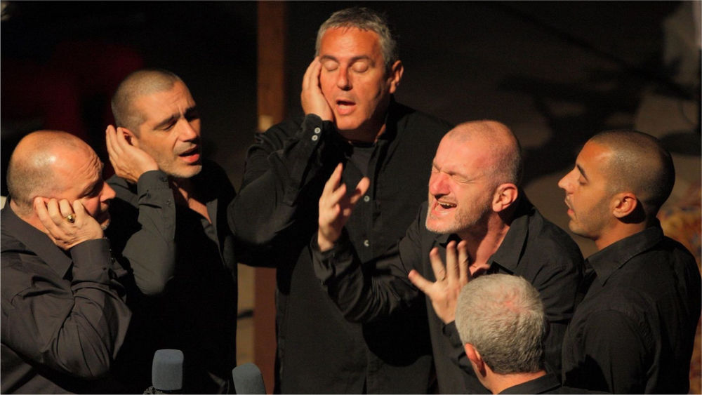 Six men wearing matching black button-up shirts stand in a semi-circle singing with eyes closed and fingers closing their ears.