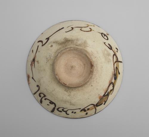 Bottom view of a circular bowl, showing the unpainted base, which is smaller than the rim’s diameter, with a raised edge. The sides are decorated in brown paint with poetry verses, over a cream background. 