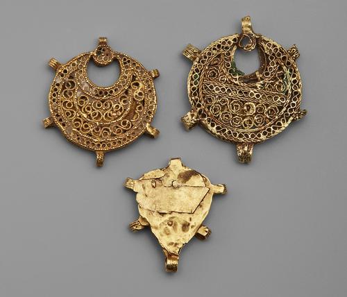 Three pendants, two closed crescent shaped and one heart shaped. All three are gold filigree and embellished with granulation. The two crescent pendants have intricate filigree on the back, while the heart shaped pendant has a solid gold back.