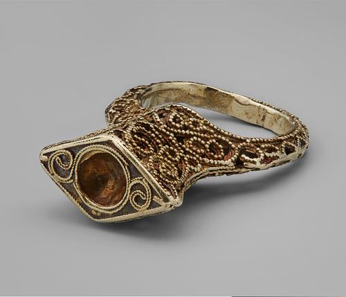 Golden ring made using filigree and granulation, laying on its side, view of the rhombus shape that rises from the bezel, featuring a round recess where a stone once was. 