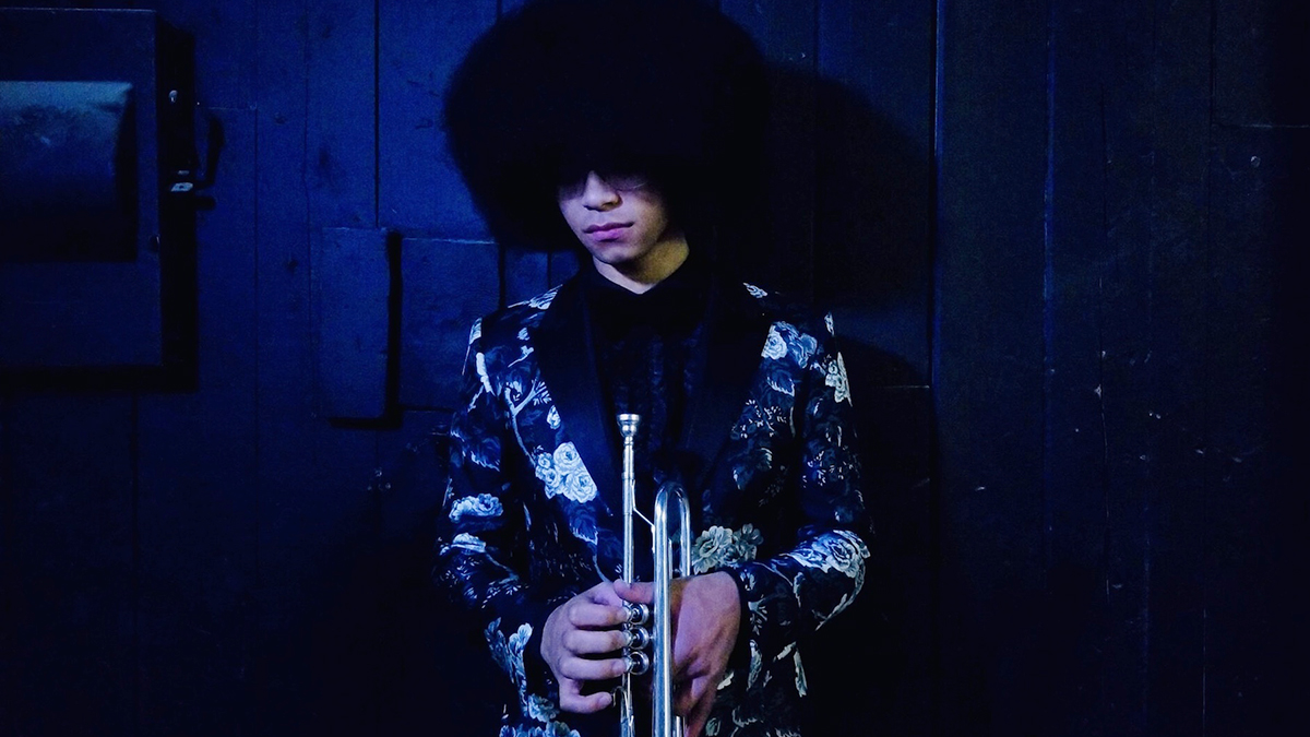 William Franklyn Leathers, wearing a floral suit jacket, holds his trumpet in a dark blue room.