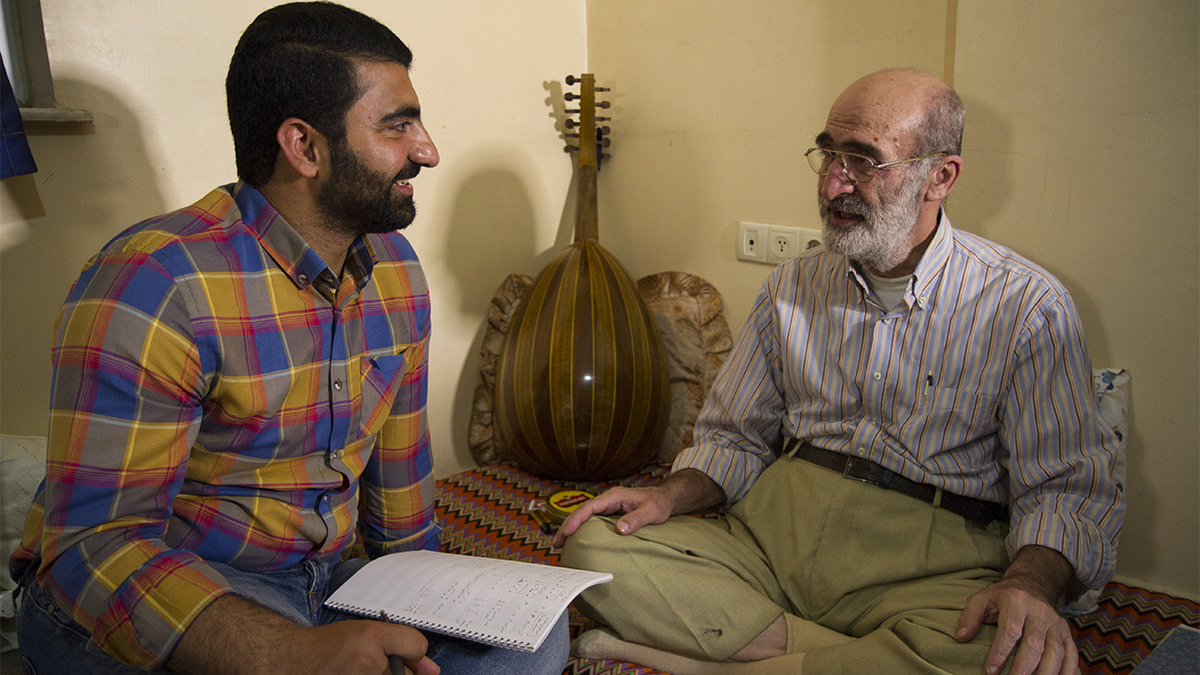 A young man and an older man sit talking, while an oud leans against the wall.