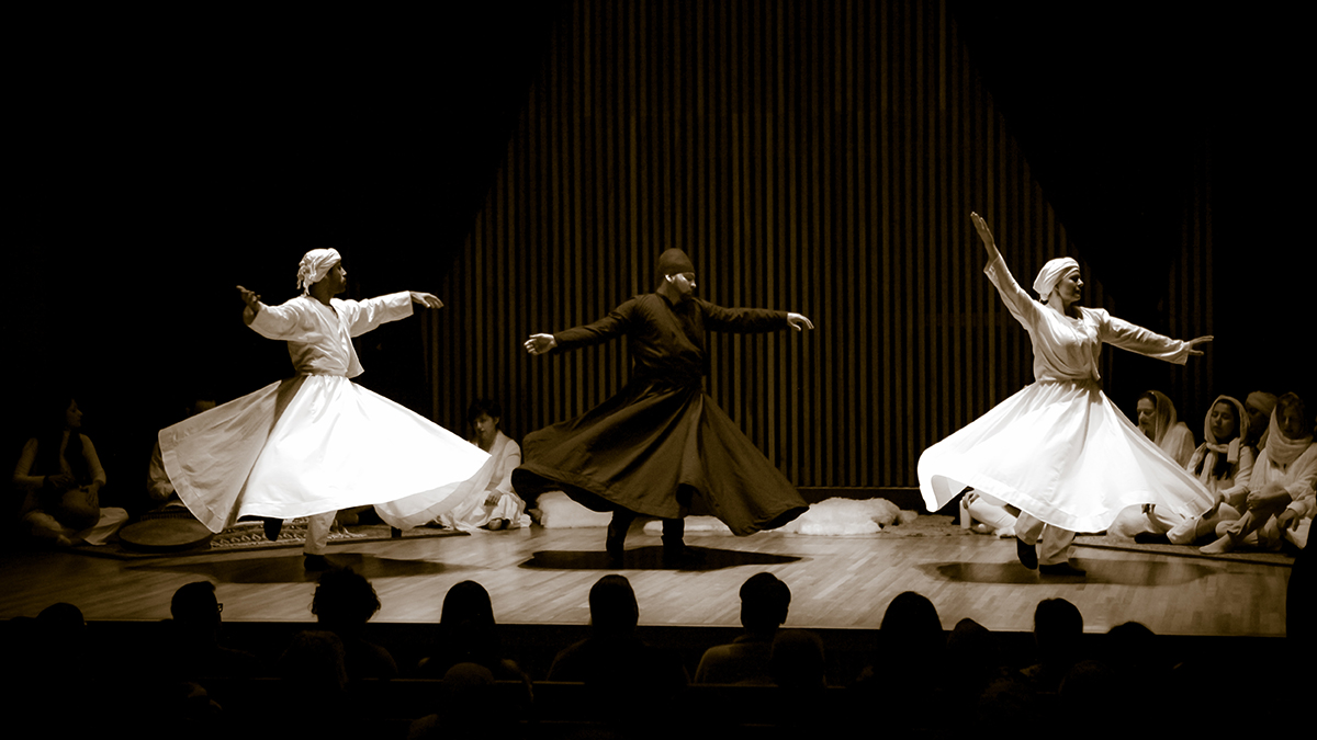 Three whirling dervishes twirl on a stage surrounded by seated onlookers.