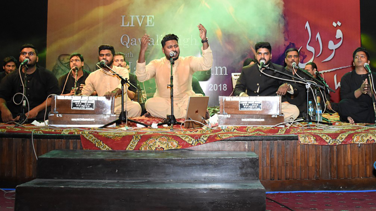 Hamza Akram and a large ensemble of musicians sit on a stage singing and playing instruments.