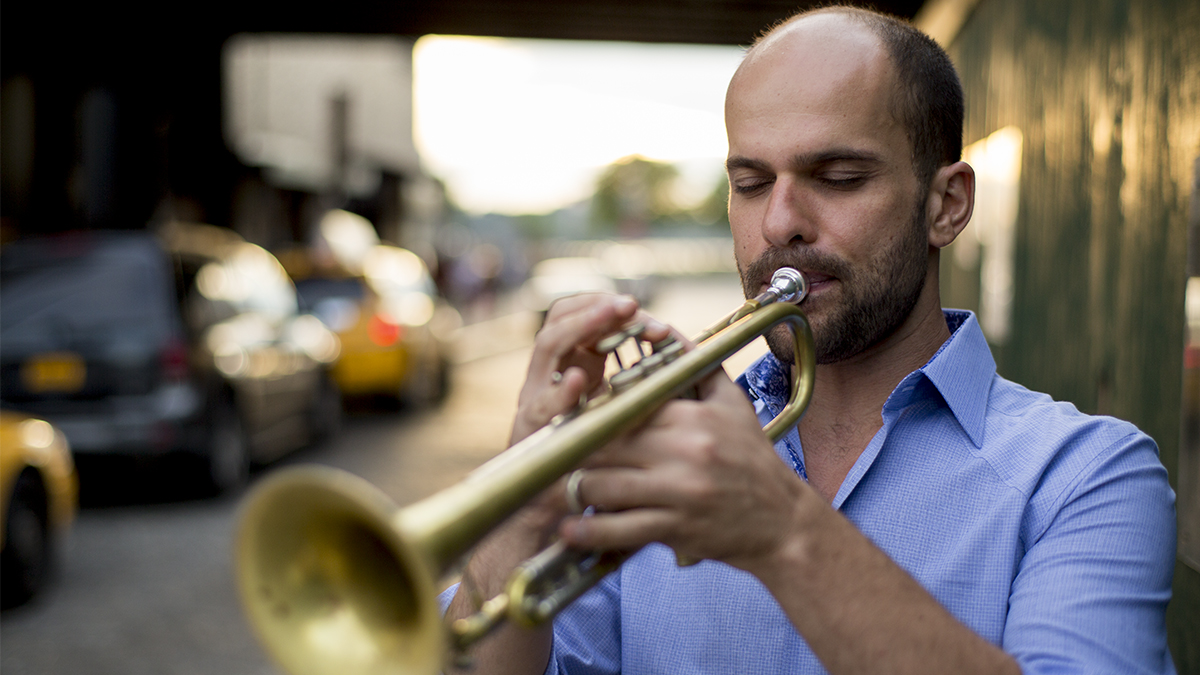 Amir ElSaffar plays the trumpet beside a street, while taxis pass by.
