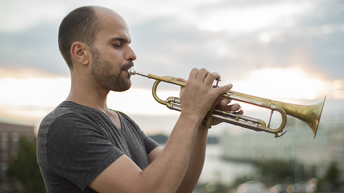 Amir ElSaffar, in profile, playing the trumpet outside, with blurry buildings and cars in the background.