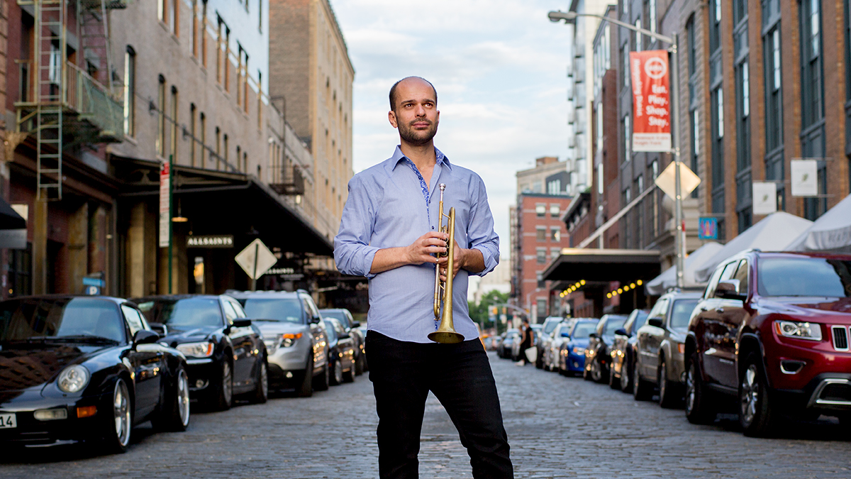Amir ElSaffar stands holding his trumpet on a street lined with parked cars and brick buildings.