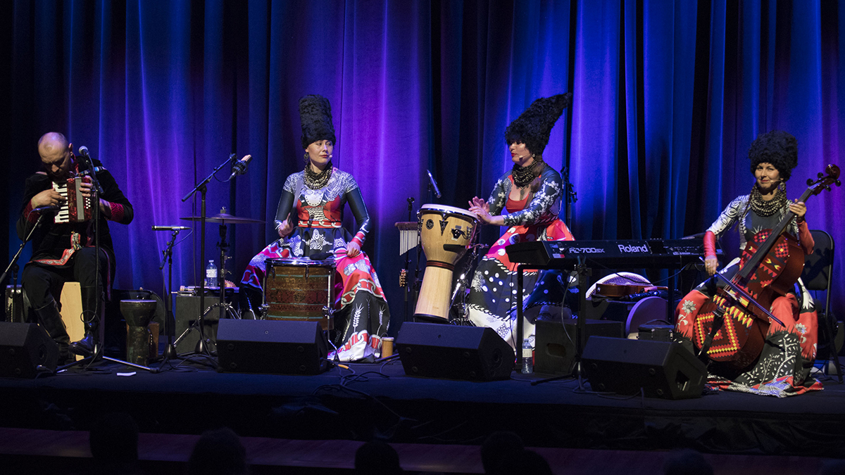 DakhaBrakha’s four members sit on chairs, in costume, playing instruments on a purple-hued stage.