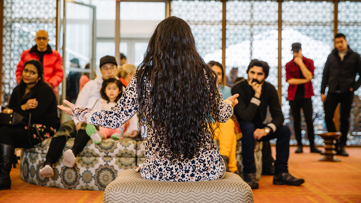 A woman wearing a white, blue, and red floral-pattered dress sitting on an ottoman with her back facing the camera and talking to a small audience of people, including families with children.