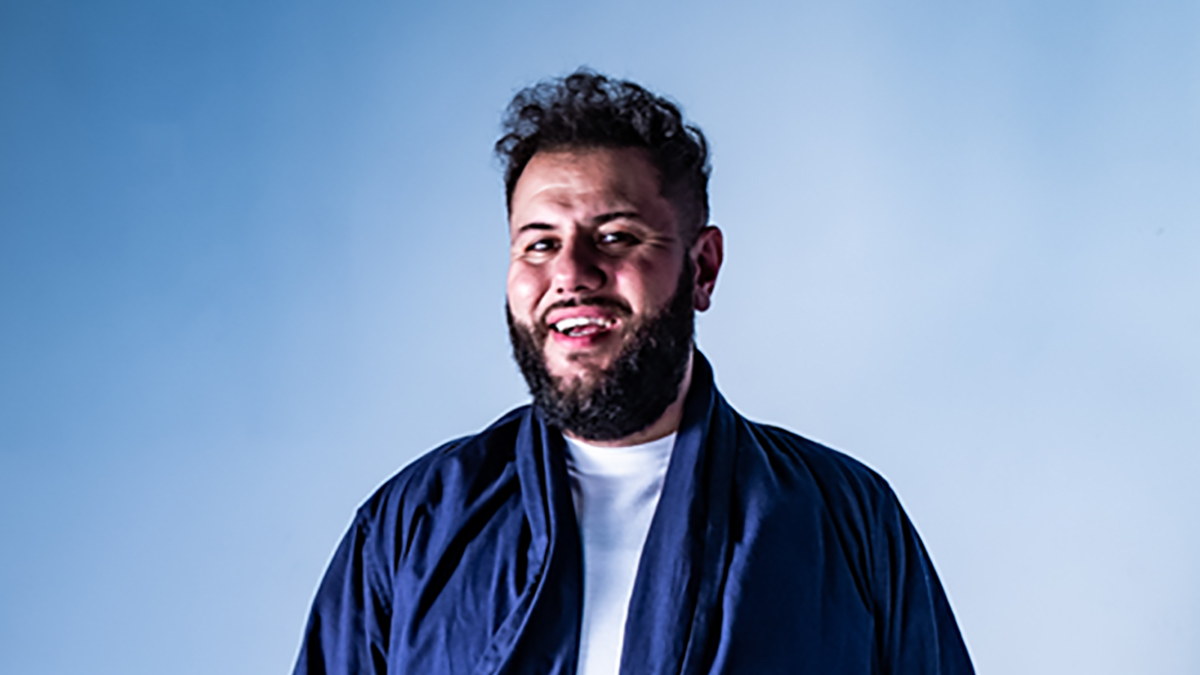 Comedian Mo Amer laughs in front of a blue backdrop
