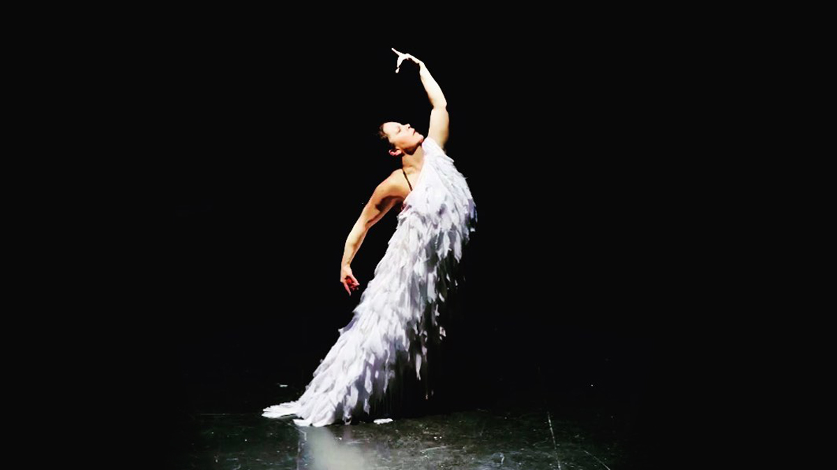Carmen Romero, wearing a purple feathered dress, strikes a flamenco pose on a dark stage.