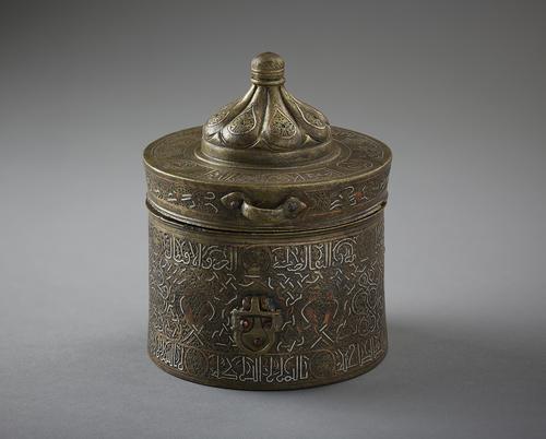 Inkwell of cylindrical form with shallow cover. Three palmette mounts one with loop handle, decorated with incised details and inlays with interlacing strap-work enclosing paired birds on an incised scroll ground, bands of inscriptions interrupted by twelve small roundels with the zodiacal signs.