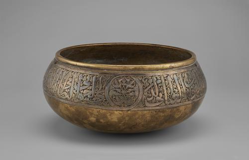 This round-bottomed brass bowl with bulging sides was raised from a single sheet of brass. View of one of three large roundels containing flying birds whose tail feathers issue from interlaced stems, placed in the Arabic inscription running around the exterior of the bowl.  