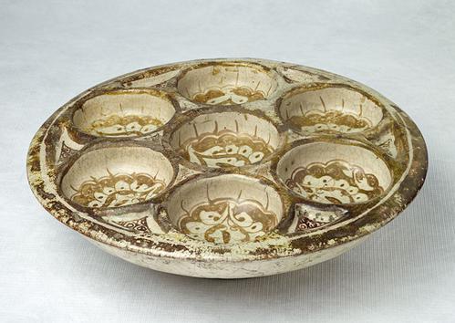 Side view of low dish contains seven hollows, six grouped around a central one. It is painted in brown lustre on a white ground.