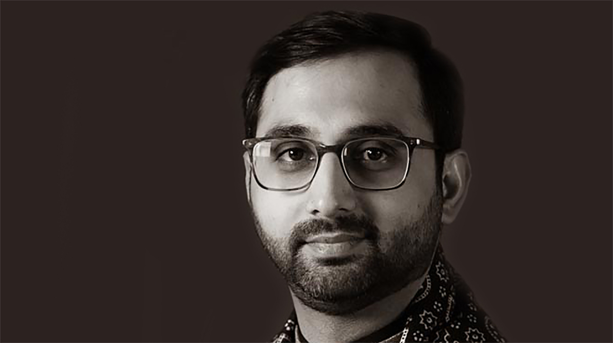 A black and white portrait of Umair Jaffar, wearing glasses and a patterned shirt. 