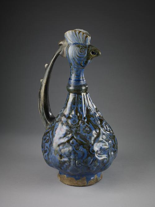A ewer made of reddish clay with blue glaze, shaped like a rooster's head. The rooster's features and its beak are painted in dark brown on top of the deep blue glaze, and handle from the top of the head to the body.