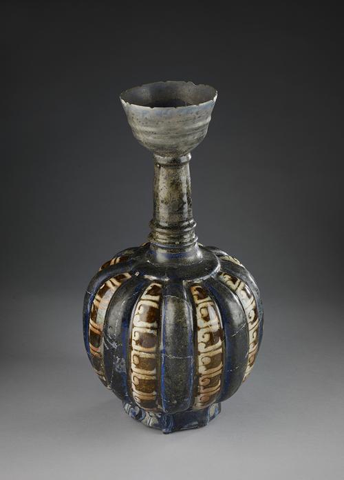 Bottle consists of a high foot, a globular body divided into sixteen projecting convex sections, a tall, thin neck and a cup-shaped mouth. Glazed dark blue except for eight of the vertical sections on the body which are decorated by white kufic letters against a dark reddish-background. 