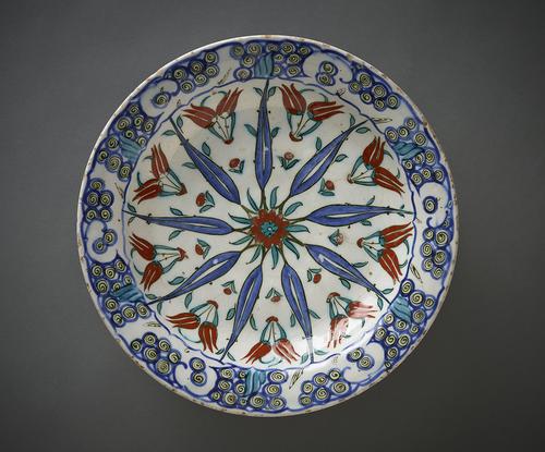 Ceramic dish, with slightly concave sides and a flat rim, the interior decorated with a radiating pattern of elongated leaves, the spaces in between with paired tulips, the rim with a series of scroll-shaped motifs, painted in red, blue, black and sage-green against an opaque-white ground.