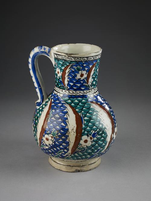 Ceramic jug, with a bulbous body, tall cylindrical neck and S-shaped handle. The body and neck decorated with a fine band of saz leaves, each conjoined with a rosette, set against a fish-scale motif, painted in green, blue, red and white with black outlines.