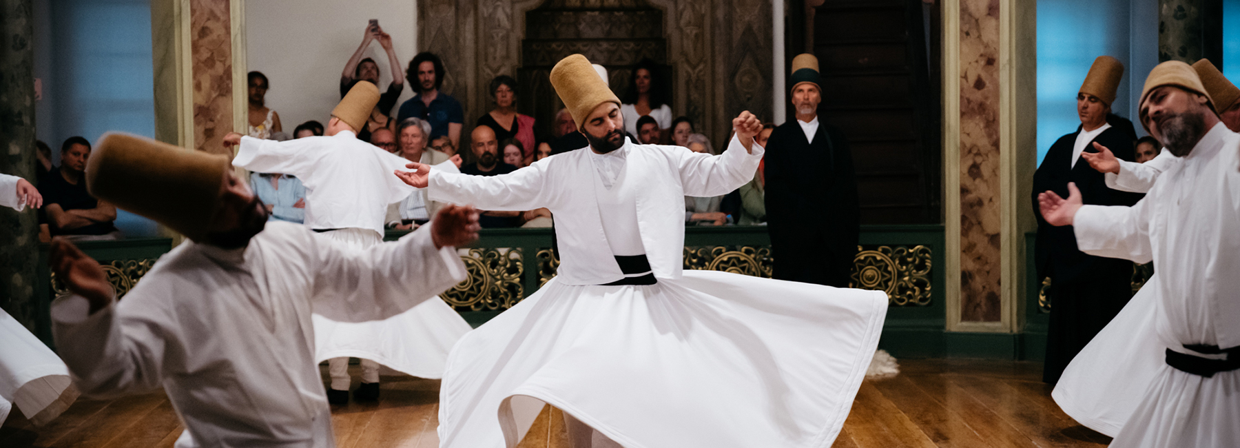 A whirling dervish in a white robe and a golden cap dances on a wooden floor while a group a spectators looks on. 