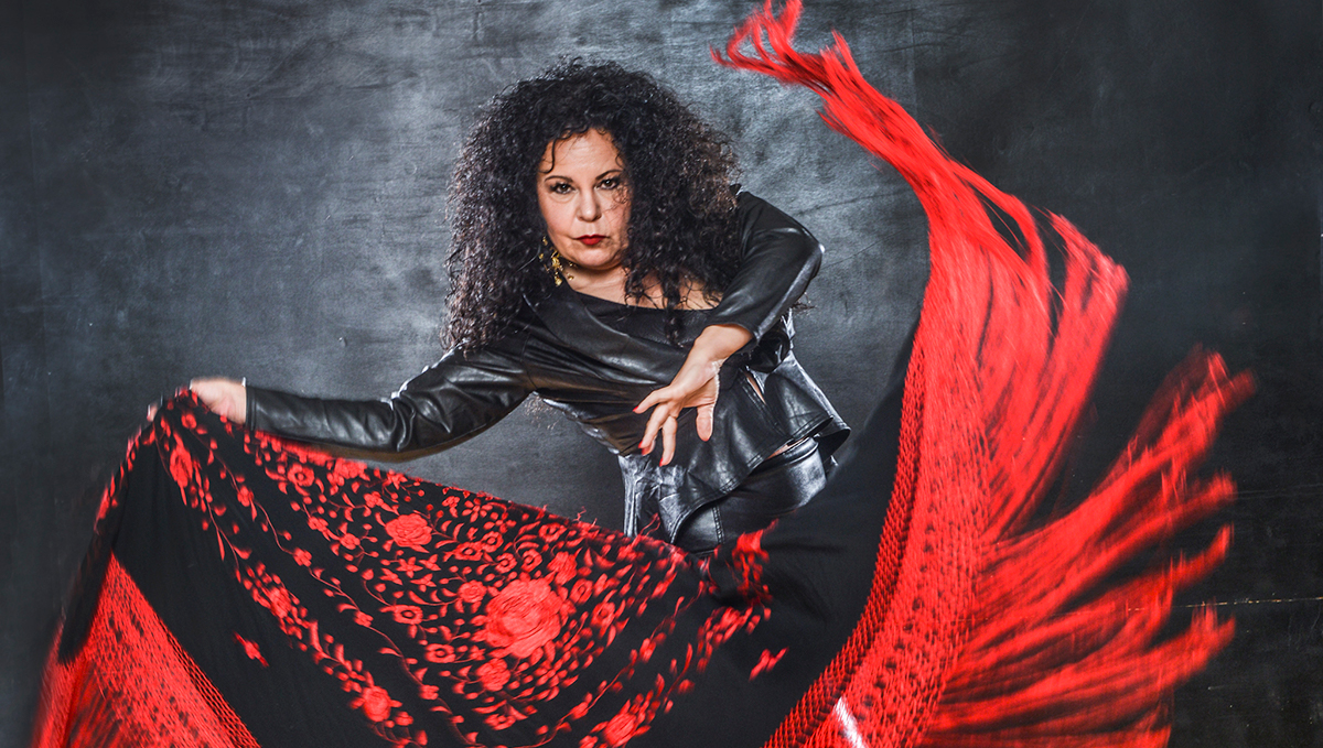 A dark-haired woman in a black leather bodysuit waves a red and black scarf as she dances. 