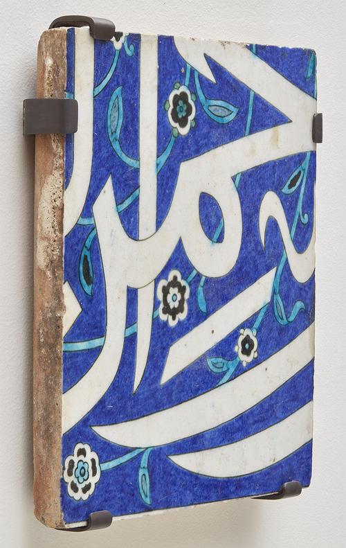 Square ceramic tile decorated with arabic calligraphy and florals, mounted on wall.