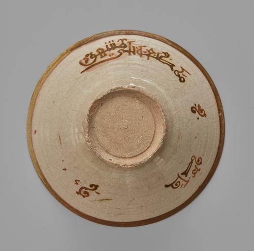 Bottom of a beige bowl with a brown plain band around the rim, the exterior with a band of angular kufic and scrolling s-motifs, and plain foot. 