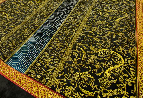 Detail of a carpet, black velvet with gold pins that create scenes of wild animals, arabesques, and long strips down the center.