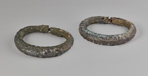 Two silver arm bracelets, engraved and feature depictions of five figures, including musicians playing a variety of instruments. Showing signs of corrosion on all sides.