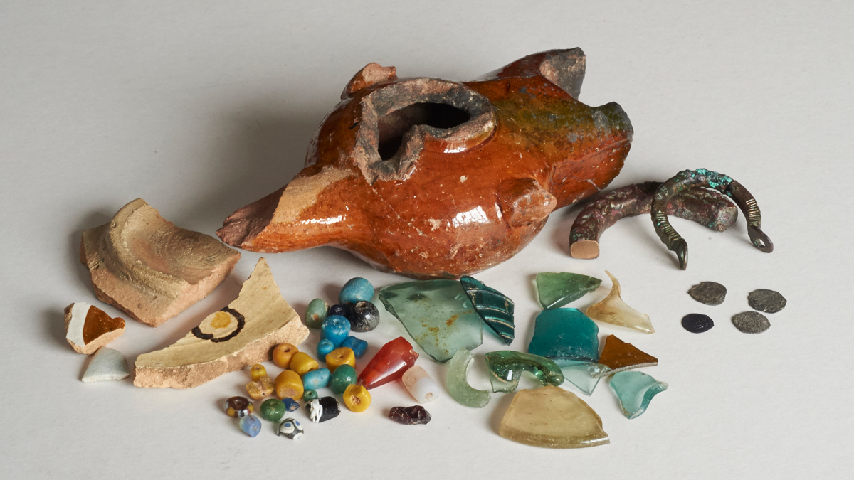 An array of fragments made of glass, ceramics, beads, coins, and metal against a grey backdrop.