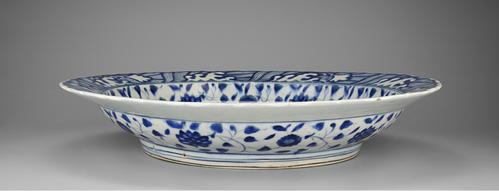 Side view of the blue and white dish that follows a Chinese model so closely that it might have passed for a Chinese original if the body had been translucent like porcelain. Blue and white designs cover the white plate with a foliage design in the centre.
