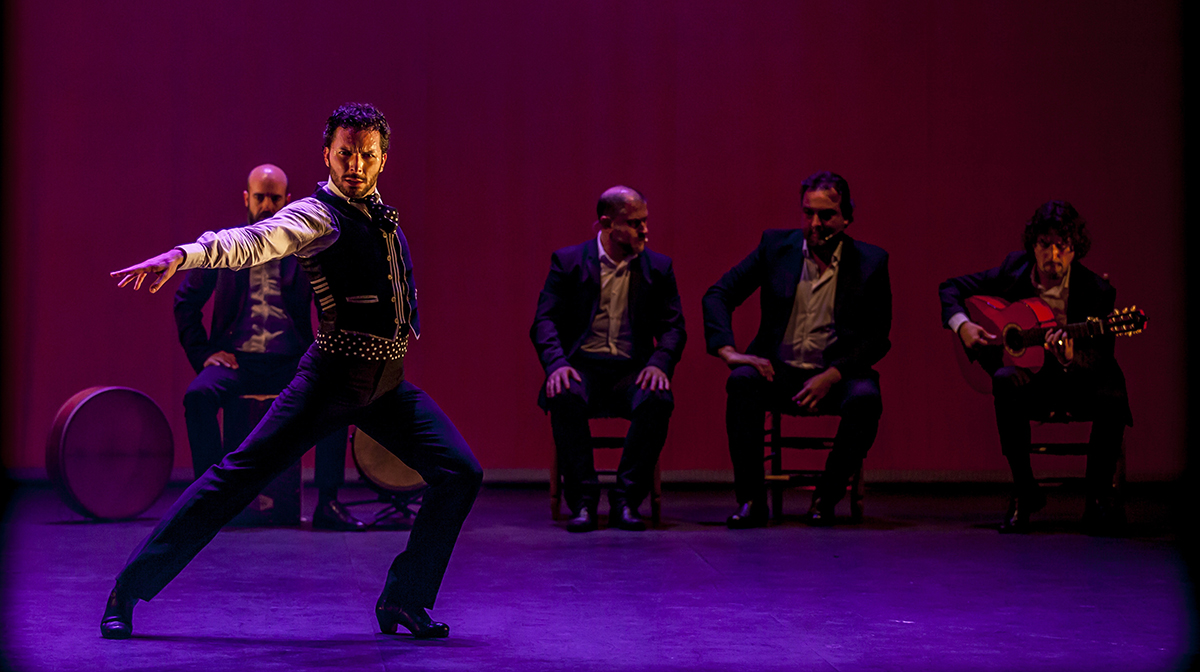 Isaac Tovar lunges on stage with a serious expression on his face, while four musicans sit upstage.
