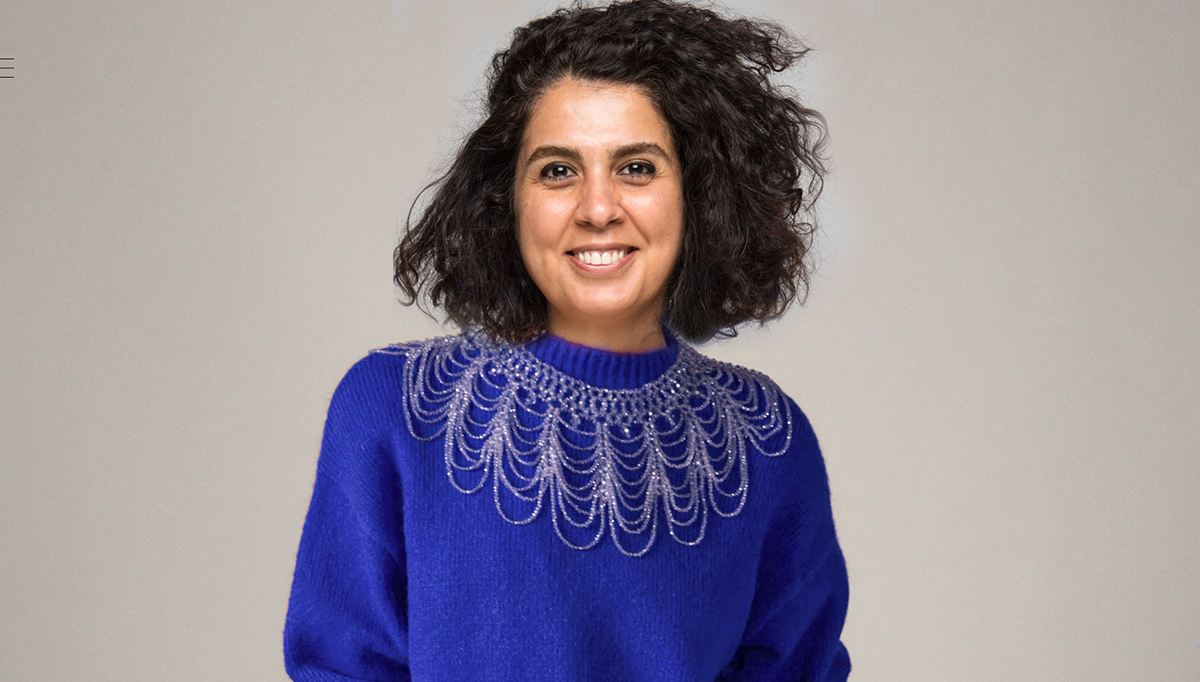 Wearing a bright blue sweater with white detailing around the collar, a woman with brown, shoulder-length curly hair smiles for the camera.