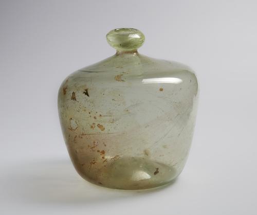 Short glass bubble shaped bottle with narrow rim, rounded mouth, blown from colourless glass with a greenish tinge. Left side of the shoulder of the bottle is higher than the right, brown speckles in the glass.
