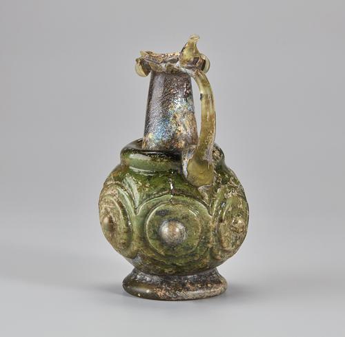 Side of a green, brown and slightly iridescent glass ewer with a cylindrical neck with an almond-shaped mouth, a circular handle connecting the mouth to the body