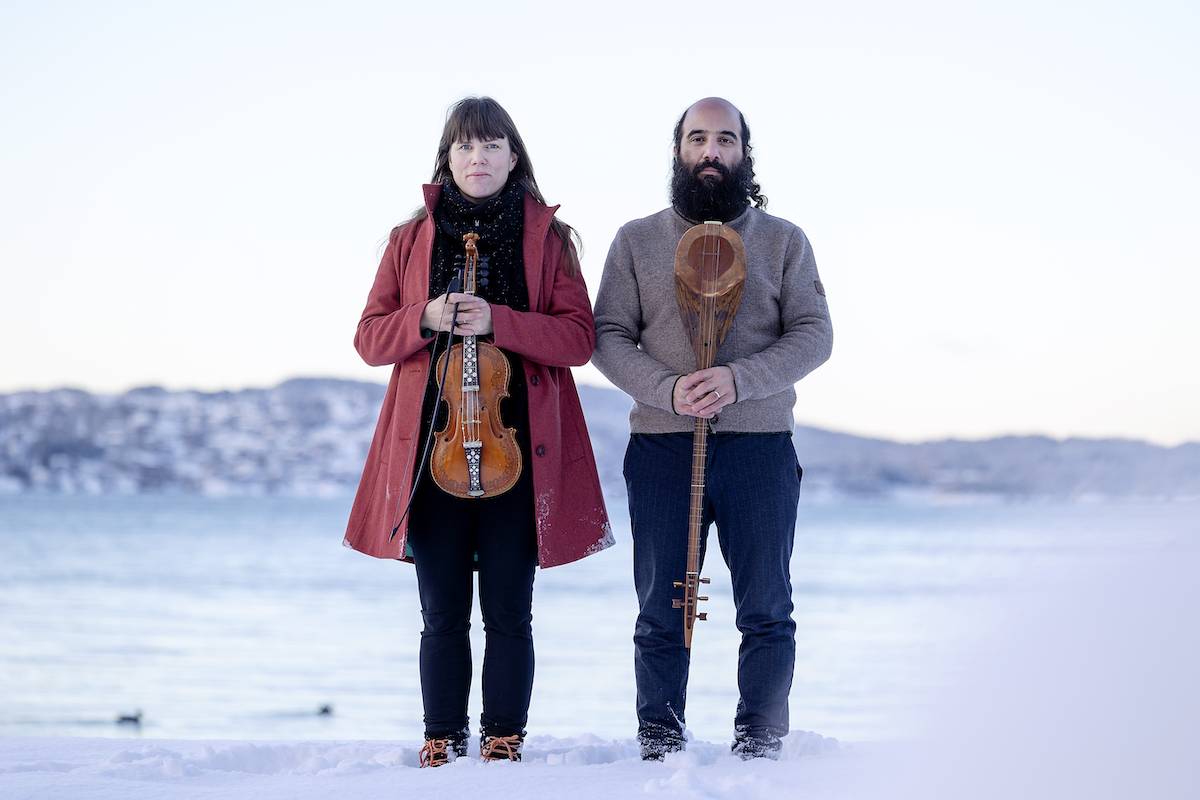 Kiya Tabassian stands in an icy terrain next to Benedicte Maurseth holding his sitar while she stands holding her fiddle. 